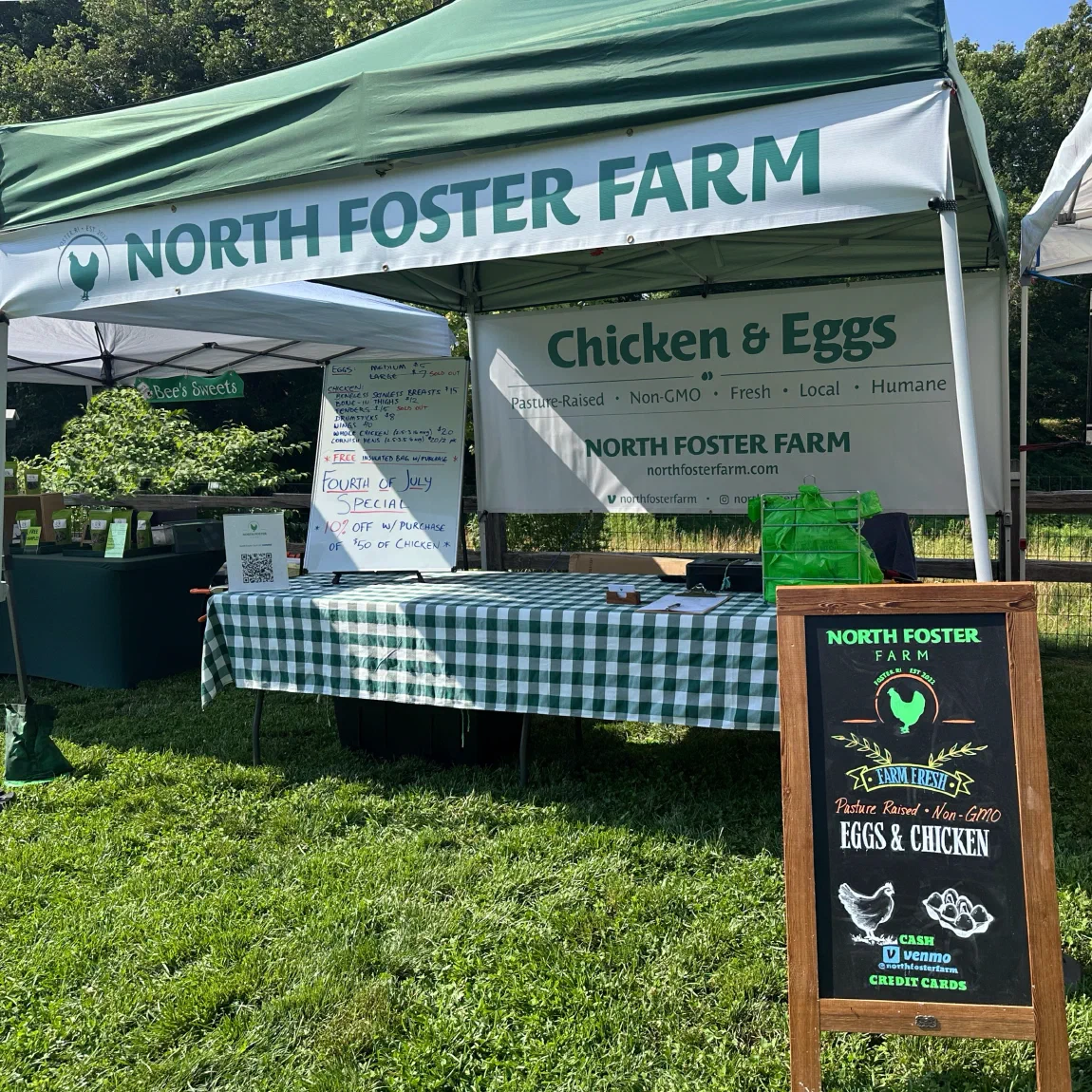 North Foster Farm's market booth on grass: a green tent over a table with a checkered tablecloth, a Chicken & Eggs banner, a whiteboard price list, and a chalkboard sign.